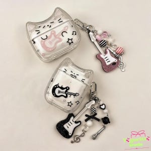 May include: Two clear AirPod cases shaped like a cat's face, each with a guitar graphic. One case has a pink guitar, the other a black guitar. Charms with guitars, beads, and musical notes dangle from the cases. The cases are on a light background.