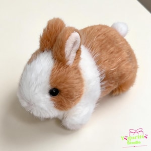 May include: A plush rabbit toy with soft brown and white fur, featuring dark round eyes, upright ears with white inner lining, and a small white tail. The bottom right corner displays the text Yguritt Studio with a small bow and cherry icon.