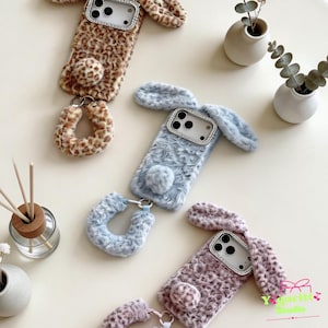 May include: Three fluffy phone cases in the shape of rabbits. One is leopard print, one is light blue, and one is a mauve color. Each case has a rhinestone-studded camera frame, bunny ears, and a fluffy tail. Matching wrist straps are also included.