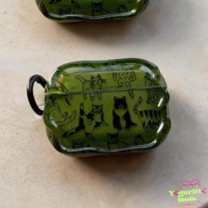 May include: Green protective case with a black handle, featuring a repeating pattern of black cat illustrations. The case is designed to hold small electronic devices. The case has a glossy finish and a rounded shape.