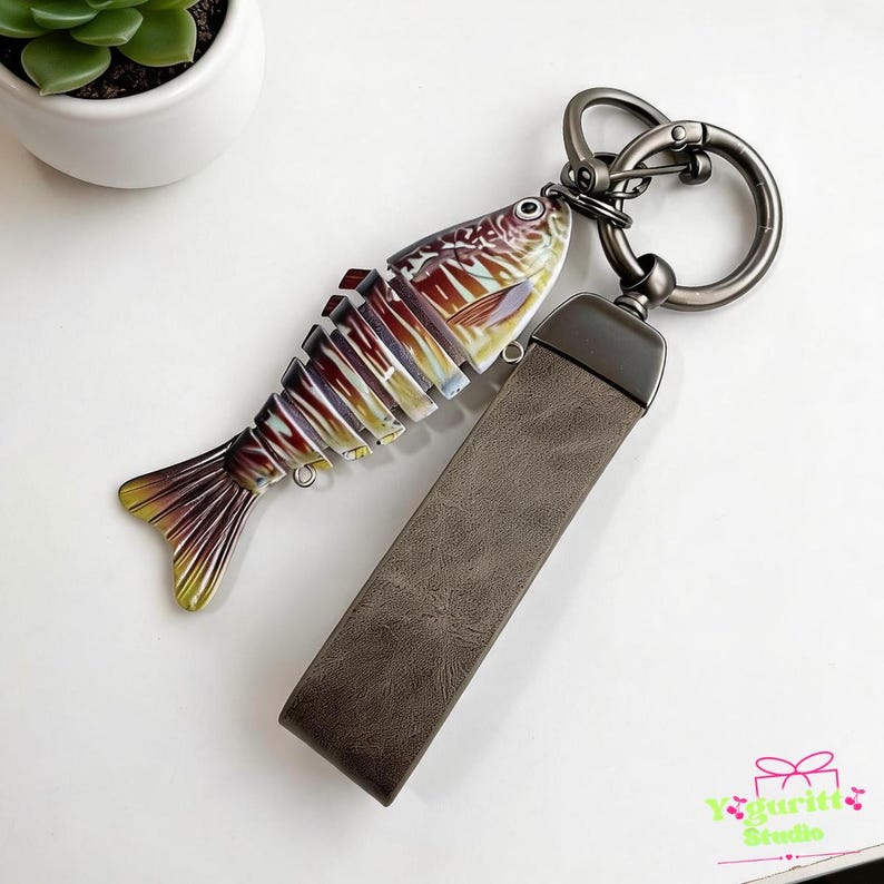 May include: A keychain featuring a multi-segmented, articulated fish charm with a metallic finish in shades of brown, gold, and cream, attached to a dark gray metal ring and a rectangular, textured dark gray faux leather strap.