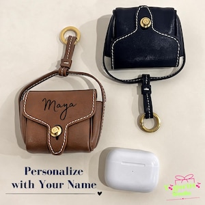 May include: Two leather cases for wireless earbuds, one brown and one black, each featuring a gold-toned snap closure, a strap with a gold ring, and white contrast stitching. The brown case is personalised with the name Maya in black script.