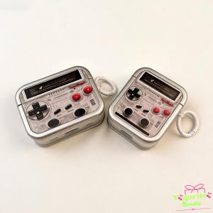 May include: Two silver protective cases for wireless earbuds, designed to resemble retro handheld gaming consoles. Each case features a black screen area, a directional pad, red and black circular buttons, and a white side-mounted attachment ring.