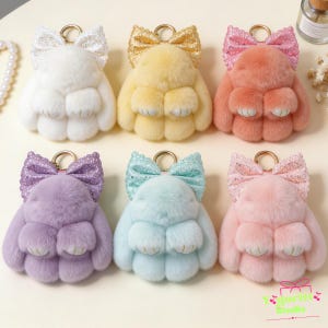 May include: Six plush rabbit keychains arranged in two rows. Each rabbit features soft fur in white, yellow, peach, purple, light blue, or pink, topped with a matching glittery bow and a gold-toned metal ring for attachment to keys or bags.