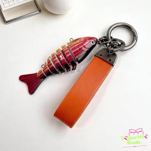 May include: A keychain featuring a segmented fish charm with red, yellow, and black stripes attached to a dark metal ring and a solid orange rectangular leather strap. The fish charm has a detailed eye and a multi-part body design.