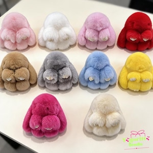 May include: A collection of ten plush rabbit keychains in various colours including pink, white, red, brown, grey, blue, yellow, magenta, and cream. Each fluffy toy features long ears and small white paws. The text Yguritt Studio is in the corner.