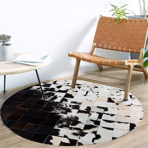 Può includere: Un tappeto rotondo in pelle di mucca con un design patchwork bianco e nero. Il tappeto è posizionato su un pavimento in legno, con una sedia in pelle e un tavolino con una pianta e libri sullo sfondo.
