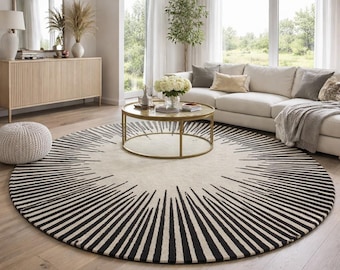 May include: A large, round area rug with a cream-colored center and a black, sunburst-like design radiating from the edge. The rug is placed on a light-colored hardwood floor in a living room setting. The room also features a white sofa, coffee table, and other decor.