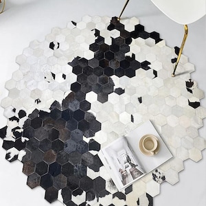 May include: Hexagon-shaped cowhide rug with a black and white pattern. The rug has a natural, organic look and feel. A white chair with gold legs is partially visible, along with a magazine and a cup on the rug.