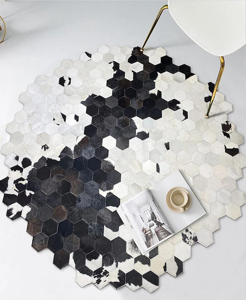 May include: Hexagon-shaped cowhide rug with a black and white pattern. The rug has a natural, organic look and feel. A white chair with gold legs is partially visible, along with a magazine and a cup on the rug.