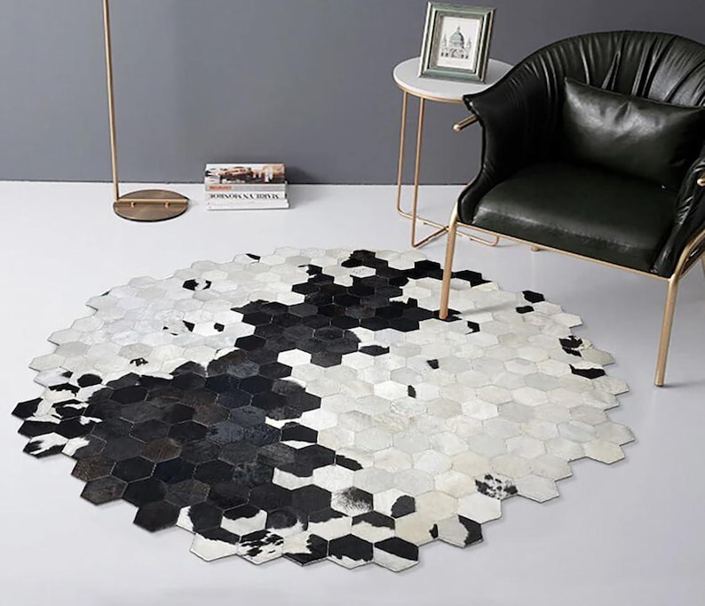May include: Hexagon-shaped rug with a cowhide pattern in black and white. The rug is placed on a white floor, with a black leather chair and a small side table in the background. The rug's design is a modern take on a classic pattern.