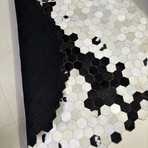 May include: A cowhide rug with a geometric pattern of black, white, and gray hexagonal shapes. The rug has a black backing and a natural, textured surface. The design creates a modern, stylish look for home decor.