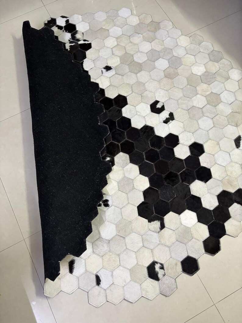 May include: A cowhide rug with a geometric pattern of black, white, and gray hexagonal shapes. The rug has a black backing and a natural, textured surface. The design creates a modern, stylish look for home decor.
