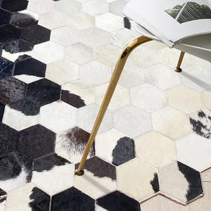 May include: A cowhide rug with a geometric hexagonal pattern. The rug features a mix of black, white, and cream-colored patches, creating a striking contrast. A gold-colored chair leg and a portion of a magazine are visible.