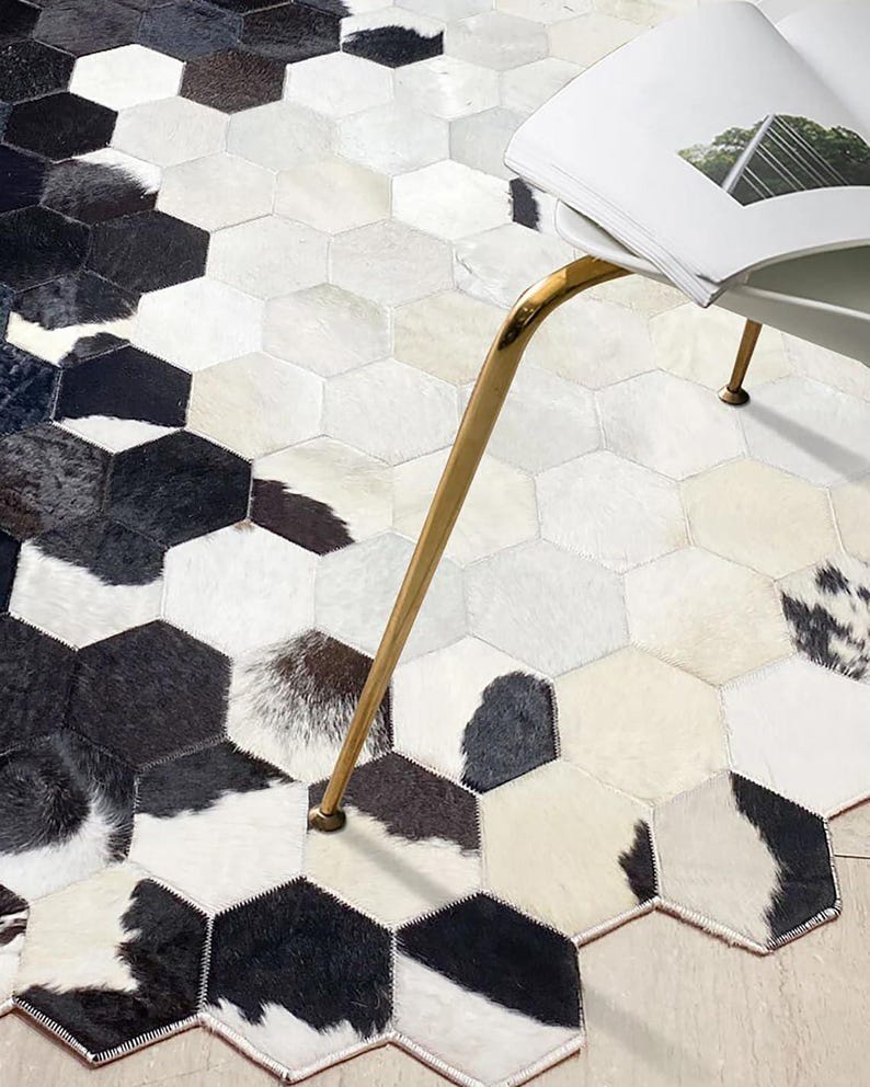 May include: A cowhide rug with a geometric hexagonal pattern. The rug features a mix of black, white, and cream-colored patches, creating a striking contrast. A gold-colored chair leg and a portion of a magazine are visible.
