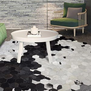 May include: A modern living room scene featuring a white round coffee table and a unique hexagonal rug. The rug has a striking black and white cowhide pattern. A green sofa and chair add color to the room.