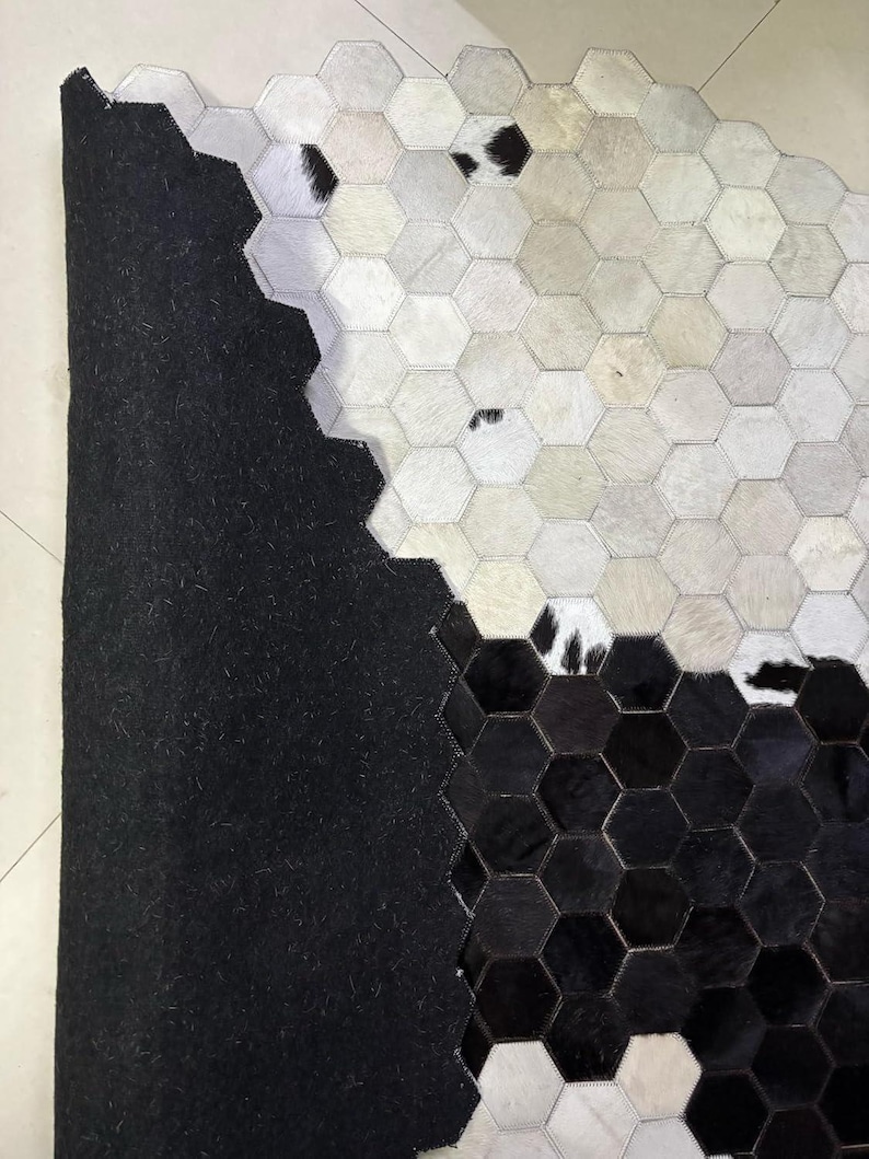 May include: A cowhide rug with a geometric pattern. The rug features a patchwork design of hexagonal shapes in black, white, and shades of gray. The rug has a black backing.