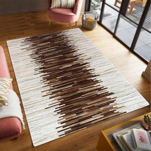 May include: Rectangular area rug with a geometric design. The rug features a central pattern of brown and tan horizontal lines against a background of off-white lines. The rug is placed on a hardwood floor.