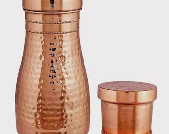 Könnte beinhalten: Eine Kupferwasserflasche und ein kleiner Kupferbehälter. Die Flasche hat eine strukturierte, gehämmerten Oberfläche und ein dekoratives Band. Die Flasche hat einen Deckel mit kleinen Löchern. Der kleine Behälter hat eine glatte Oberfläche und einen Deckel.