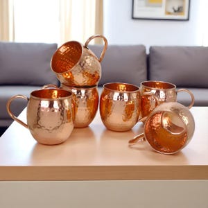 Puede incluir: Un conjunto de tazas Moscow Mule de color cobre con texturas martilladas. Las tazas tienen cuerpos redondeados, asas curvas y están dispuestas sobre una superficie clara. El color cobre refleja la luz, creando un ambiente cálido.