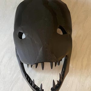 Può includere: Una maschera nera stampata in 3D con un design minaccioso. La maschera presenta fori per gli occhi, un naso a punta e una bocca spalancata con denti affilati. La maschera è probabilmente per cosplay o costumi.