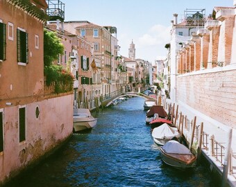May include: A scenic canal in Venice, Italy, with boats docked along the sides. Buildings with orange and beige facades line the waterway, and a bridge spans the canal. The sky is clear blue.
