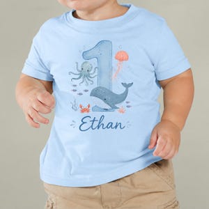 May include: A light blue short-sleeved t-shirt featuring a large blue number one graphic decorated with watercolor-style illustrations of an octopus, a whale, a jellyfish, and a crab, with the name Ethan printed in dark blue script below.