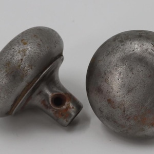 May include: Two antique metal door knobs. One knob is round, the other is oval with a stem. Both knobs have a weathered, aged appearance with rust-colored spots, suggesting they are vintage or reclaimed.