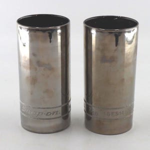 May include: Two cylindrical metal sockets with a dark, weathered finish. Each socket features the engraved text Snap-on and SFS141 on the lower band. The tools have a hollow interior and a smooth, reflective surface showing signs of use and oxidation.