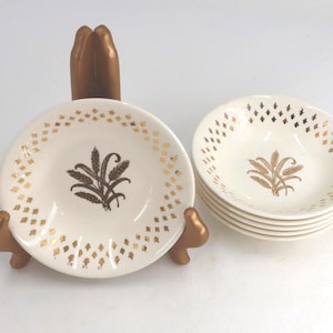 May include: A set of small white ceramic bowls featuring a gold wheat stalk design in the center and a gold diamond pattern along the rim. One bowl is displayed on a gold-colored stand, while the others are stacked neatly to the right side.