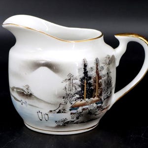May include: White ceramic creamer with a landscape design. The creamer features a curved spout, a handle with gold trim, and a painted scene of trees, mountains, and water. The rim and handle are accented with gold. A decorative piece for serving.