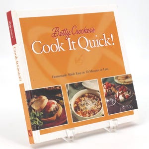 May include: A cookbook titled "Betty Crocker's Cook It Quick!" is displayed on a clear plastic stand. The cover is orange with the title in white and red. The book features images of prepared meals, including a burger, soup, and dessert.