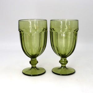 May include: Two olive green water goblets. The glasses have a ribbed design on the bowl and a detailed stem. The base is round and the glass has a vintage aesthetic. The glasses are made of glass.