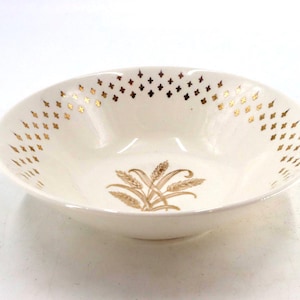 May include: A round, off-white ceramic bowl featuring a central illustration of golden wheat stalks. The interior rim is decorated with a repeating pattern of small, metallic gold fleur-de-lis symbols arranged in a horizontal band around the bowl.