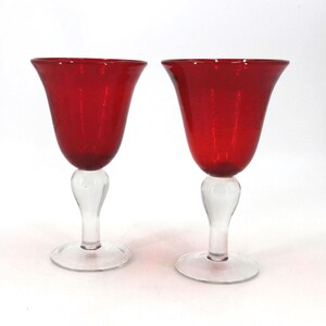 May include: Two matching goblets featuring deep red glass bowls with a subtle textured finish. Each goblet is supported by a clear glass stem and a circular clear glass base, creating a distinct contrast between the red and clear glass elements.