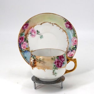 May include: A vintage teacup and saucer set. The white porcelain cup and saucer are adorned with hand-painted pink roses and gold trim. The cup is on a small metal stand. The saucer has a scalloped edge.
