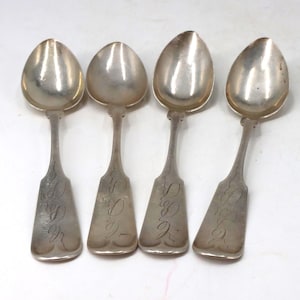 May include: A set of four silver-toned metal spoons arranged in a row. Each spoon features a rounded bowl and a flat, flared handle decorated with an intricate, engraved script monogram. The metal surfaces exhibit a polished, reflective finish.