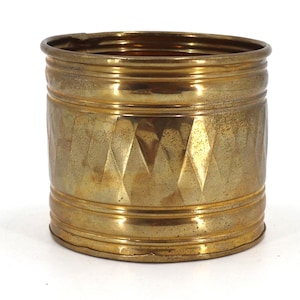 May include: A cylindrical brass container featuring a polished metallic finish with a textured diamond pattern around the center. The vessel has horizontal ridged bands at the top and bottom edges, showcasing a warm, golden-toned surface.