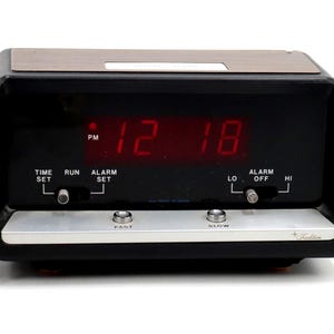 May include: Black and silver digital alarm clock with a wood-grain top. The red LED display shows the time as 12:18 PM. Features include time set, alarm set, and fast/slow buttons. A black power cord is attached.