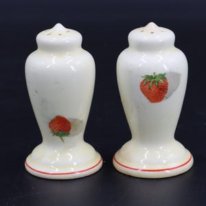 May include: A pair of cream-colored ceramic salt and pepper shakers. Each shaker features a red strawberry design and a red rim at the base. The shakers have a classic, vintage design.