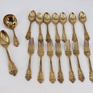 May include: A set of gold-toned flatware including seven spoons, six forks, and one ladle. Each piece features an ornate, textured handle with intricate detailing. The collection is arranged on a plain surface, showcasing the polished metallic finish.