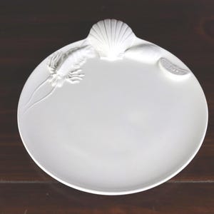 May include: A white, round serving plate with embossed seashell and marine life details. The plate features a seashell handle and is decorated with a lobster and a slice of lemon. The plate is made of ceramic and is suitable for serving appetizers or small dishes.