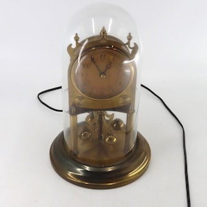 May include: A brass torsion clock with a circular face, black hands, and Arabic numerals, enclosed within a clear glass dome. The clock features a rotating pendulum with four small brass spheres and is connected to a thin black electrical power cord.