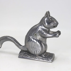 May include: A silver-colored metal nutcracker shaped like a squirrel sitting on a log base. The squirrel has a curved tail, defined facial features, and a visible metal screw at the joint where the lever mechanism connects to the body.