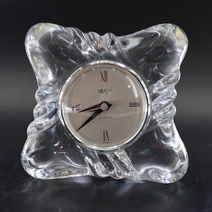 May include: A clear glass desk clock with a square, wavy-edged frame. The circular clock face features a silver-toned background, black Roman numerals at the twelve, three, six, and nine o'clock positions, black hands, and the text Mikasa Quartz.