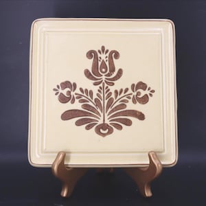 May include: A square, cream-colored ceramic plate featuring a brown, symmetrical floral pattern in the center. The plate has a raised border and is displayed on a small, curved, metallic-toned stand that supports the bottom edge.