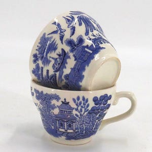 May include: Two stacked teacups with a blue and white Willow pattern. The cups are made of porcelain and have a classic design with a handle. The blue pattern depicts a landscape scene.