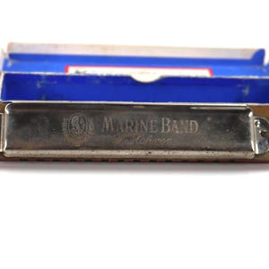 May include: A vintage Marine Band harmonica with a silver metal cover and wooden frame, resting in its blue and red box. The harmonica has the words "MARINE BAND" and "Made in Germany" printed on it.