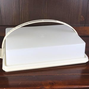 May include: A rectangular, off-white cake carrier with a matching arched handle. The carrier has a lid and a base, and is made of a durable plastic material. The handle is attached to the lid with two small brackets.