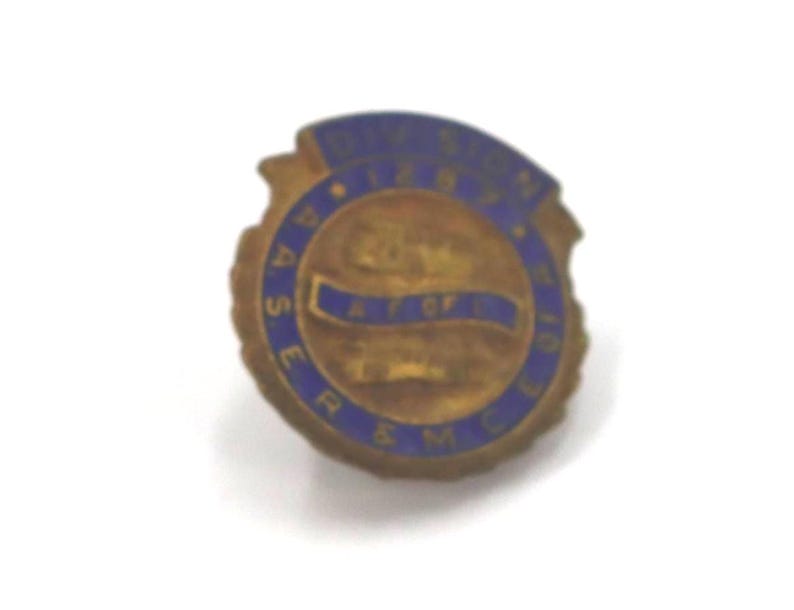 May include: A vintage brass-toned pin with blue lettering and accents. The pin features the text "DIVISION 10" at the top and "A.S.E.R. & M.C." around the outer edge. A blue banner is in the center.